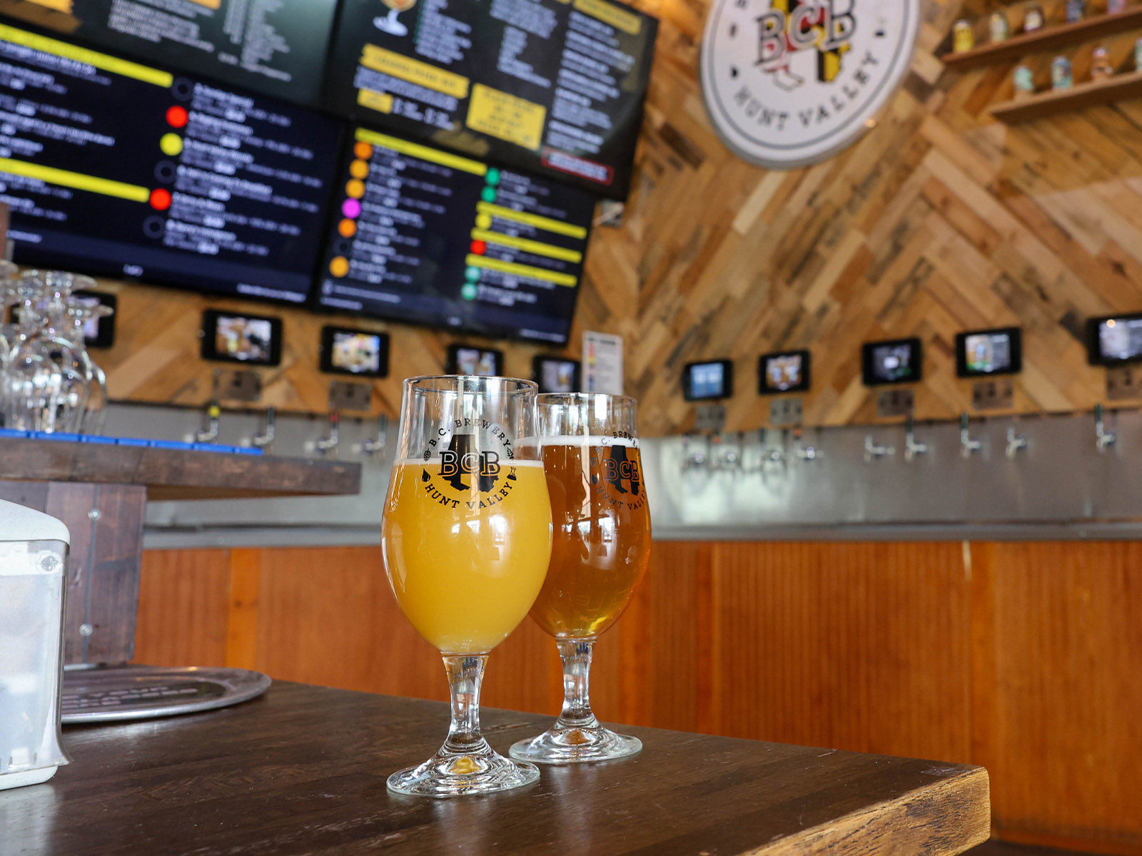 Two glasses of golden and amber craft beer on a wooden counter in front of the self-serve beer tap wall at B.C. Brewery in Hunt Valley, Maryland.