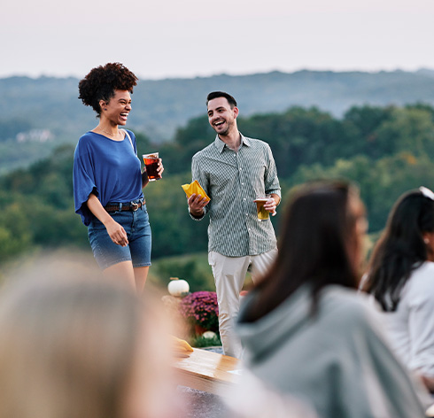 A couple laughing while playing cornhole with beers in hand at a brewery in Baltimore County with a beautiful overlook view of rolling hills.