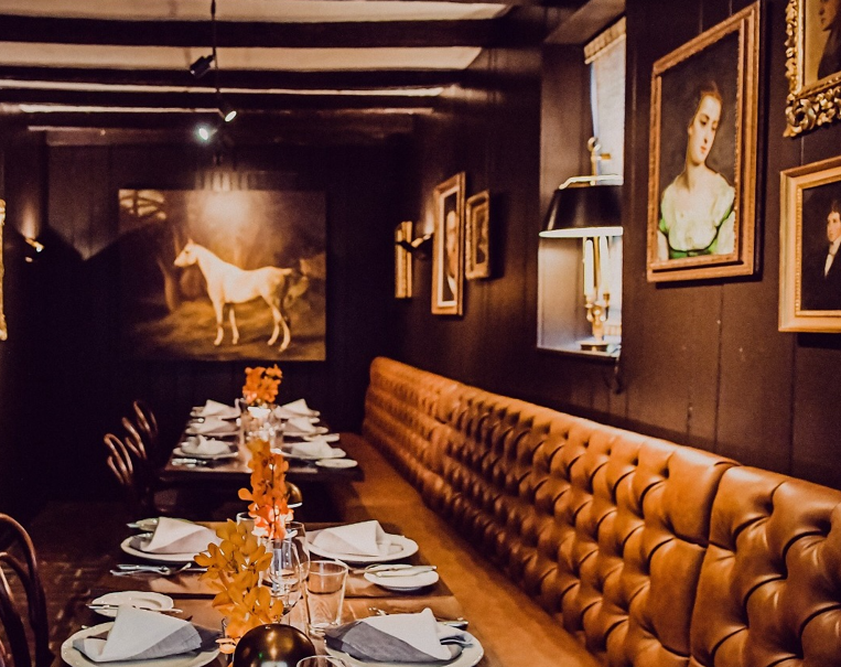 A moody image of a restaurant featuring a beautifully placed long table with bench seating and images of horses, along with portraits of various people on the wall.