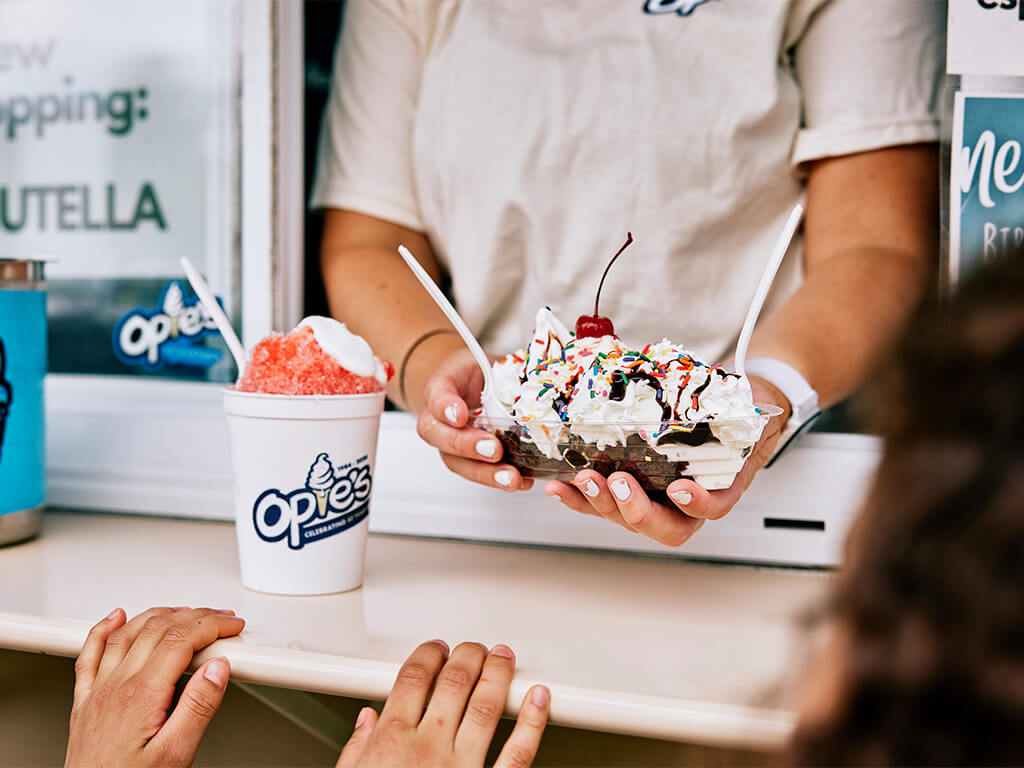 Opie’s serves a whipped-cream-topped sundae with sprinkles and a cherry at a snowball stand, with a red snowball nearby and a child waiting at the counter.